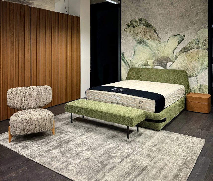 Moderní ložnice s elegantní postelí v zeleném čalounění a botanickým motivem na tapetě. Křeslo s tkaným potahem a dřevěný stolek dotvářejí útulnou atmosféru.